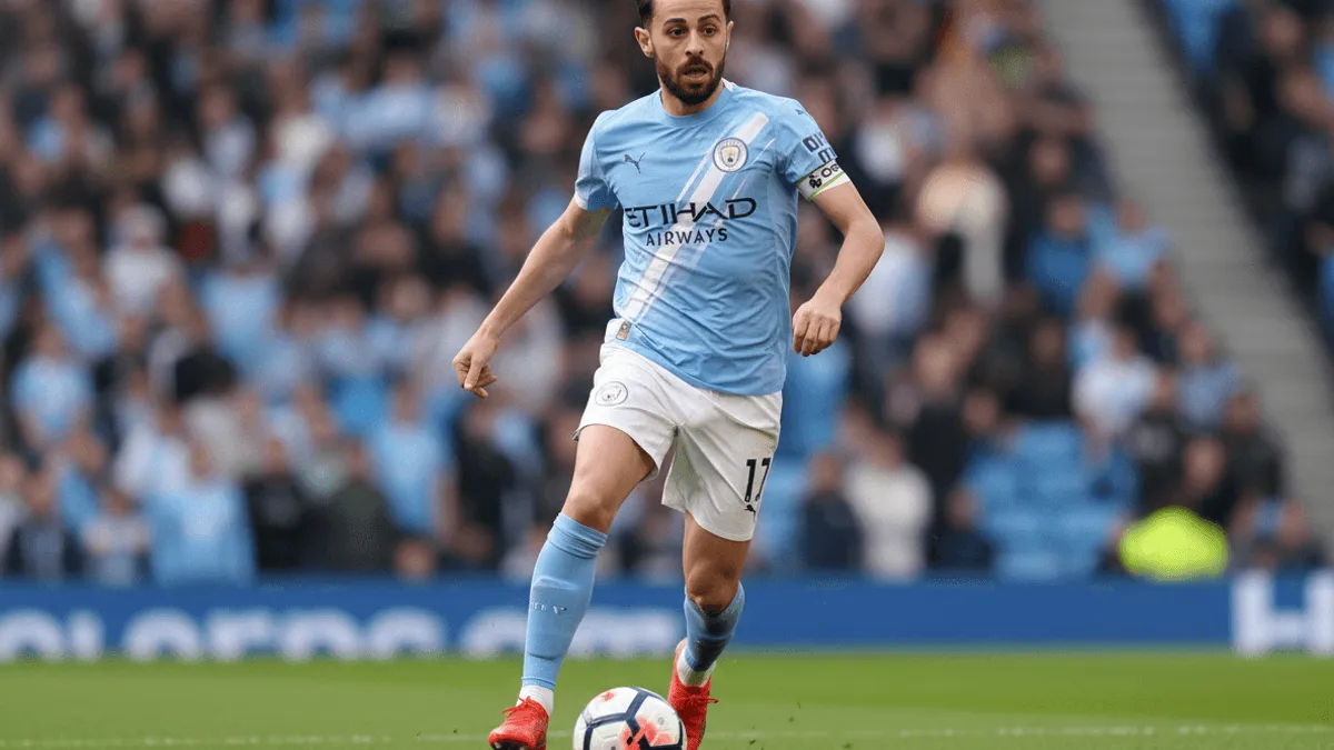 A male soccer player in a light blue jersey dribbles a ball on a green field during a match with spectators in the background.