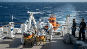 A group of medical personnel in hazmat suits prepare to transfer two patients on stretchers from a cruise ship to a smaller rescue boat. Hantavirus