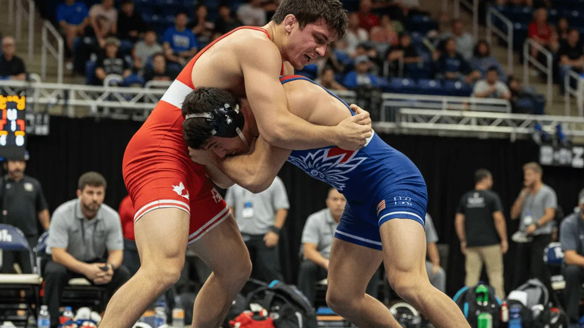 Two male wrestlers, one in a red singlet and one in a blue singlet, locked in a struggle on a blue mat.